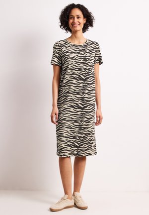Femme souriante aux cheveux bouclés portant une robe imprimé zèbre courte aux genoux avec des manches courtes et des baskets beige clair, debout devant un fond uni.