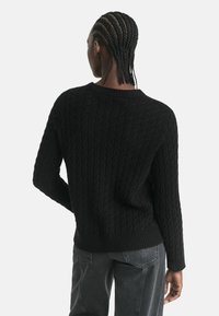 Schwarzer, gestrickter Pullover mit einem strukturierten Zopfmuster, mit einem Rundhalsausschnitt und einem gerippten Saum, kombiniert mit grauen Jeans.