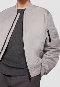 Personne portant une veste bomber gris clair avec une poche zippée et une étiquette Calvin Klein sur la manche, un pull côtelé gris foncé en dessous.