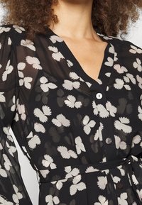 Černé průsvitné šaty s květinovým vzorem v jemných barvách, výstřihem do V, zapínáním na knoflíky a detailem stahování v pase.