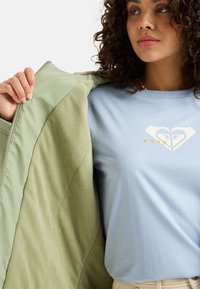 Zelená zipová bunda s fleecovou podšívkou, pod ktorou sa objavuje svetlomodré tričko s krémovým a zlatým srdcovým logom.