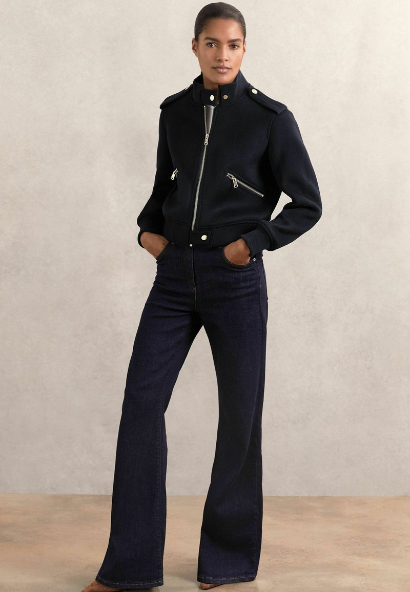 Femme portant une veste noire zippée avec des zips argentés et un jean bleu foncé évasé, se tenant les mains dans les poches devant un fond neutre.