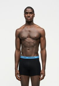 Μυώδης άνδρας με σκούρο δέρμα που στέκεται με το πρόσωπο στραμμένο προς τα εμπρός, φορώντας μαύρο μπόξερ με μπλε μέση που γράφει «BJÖRN BORG» σε απλό ανοιχτό φόντο.