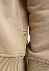 Beigen värinen collegepaitakangas, jossa on resorit hihansuissa ja helmassa. Sivusaumassa pieni oranssi lippu, jossa musta logo. Pehmeä pinta.