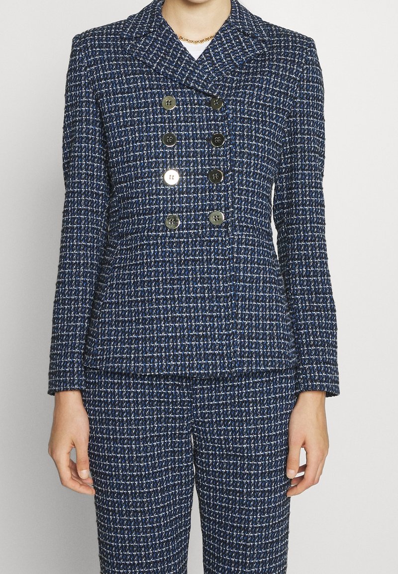 Femme portant un blazer croisé à carreaux bleu marine et blanc avec un pantalon assorti et un collier chaîne en or.