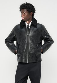Veste en cuir noir avec une finition texturée et un col en fourrure. Fermeture éclair argentée et poches latérales; coupe décontractée avec des coutures visibles.