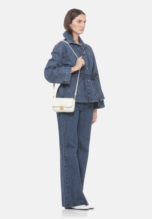 Donna in piedi che indossa una giacca di jeans con cintura e jeans a gamba larga abbinati, tiene una borsa a tracolla bianca con chiusura dorata, scarpe bianche.