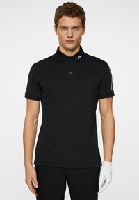 Svart polo t-shirt med krage, tre knappslå och korta ärmar. Har vit logotyp på vänster axel. Slät textur.