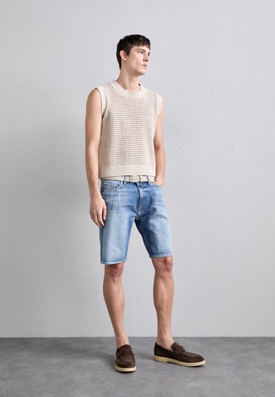 Beige gebreide mouwloze top, lichtblauwe denim shorts, bruine suède schoenen met lichte zolen, staande tegen een plain grijze achtergrond.