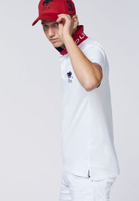 Weißes Poloshirt mit rotem Kragen und Akzenten, das ein blaues Logo auf der Brust zeigt. Das Model trägt eine rote Kappe mit einem Emblem und der Nummer 3.