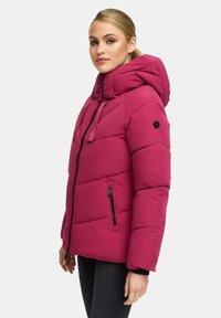 Vrouw met blond haar in een paardenstaart, gekleed in een felle roze gewatteerde jas met hoge kraag en capuchon, staand tegen een witte achtergrond.