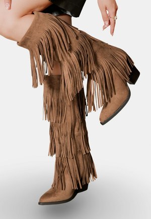 Bottes en suède marron au-dessus du genou avec des détails en franges superposées, un bout pointu et un petit talon empilé. Surface texturée avec fermeture éclair.