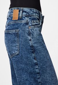 Vidutinio plačiainimo džinsai su šiek tiek išblukusiu paviršiumi, turintys rudą odinį etiketę, klasikinį penkių kišenių dizainą ir tiesų kirpimą.