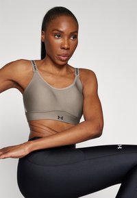 Beige sportsbh med justerbare stropper som har Under Armour-logo, kombinert med svarte tights. Glatt tekstur og atletisk design.
