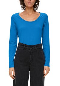 Blaues langärmliges Oberteil mit weitem Rundhalsausschnitt, aus glattem Stoff, kombiniert mit hochgeschnittenen schwarzen Jeans mit doppeltem Knopfverschluss.