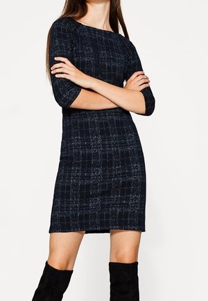 Vrouw met lang haar in een zwarte en blauwe jurk met textuur tot op de knieën en zwarte suède laarzen tot net onder de knie, armen gekruist tegen een witte achtergrond.