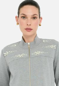 Giacca grigia con zip, caratterizzata da dettagli di perle sulle spalle e sul petto, con colletto alto e tessuto liscio e texturizzato.