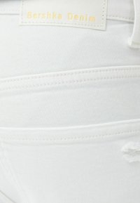 Jeans de mezclilla blanca con costuras visibles, un detalle desgastado y una etiqueta de marca etiquetada como "Bershka Denim" en texto amarillo en relieve.