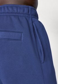 Nærbilde av en person som har på seg blå joggebukser med elastisk midje, hvor både sidelommene og baklommen er synlige på høyre hofte.