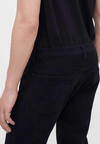 Personne portant un jean noir et une chemise noire, montrant la poche arrière et le côté du jean sur un fond uni clair.