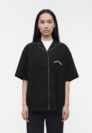 Kobieta o długich czarnych włosach, ubrana w luźną czarną koszulę zapinaną na guziki z białym obszyciem i napisem „palm angels” na kieszeni na piersi.