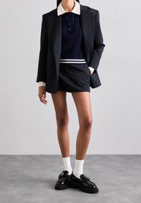 Marinblå kavaj med kontrasterande vit krage, marinblå kabelstickad tröja, skräddarsydda shorts, samt glansiga svarta loafers med detaljer i guldspänne.