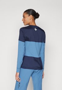 Ilgi piedurkņu sporta krekls ar tumši ziliem un gaiši ziliem paneļiem, faktūras audums un logo detalizācija aizmugurē. Standarta piegriezums ar apaļu kakla izgriezumu.