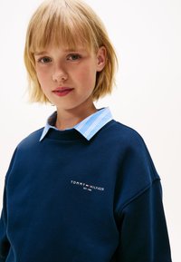 Blonde kind met groene ogen in een blauwe Tommy Hilfiger sweatshirt over een lichtblauw gestreept overhemd tegen een plain witte achtergrond.