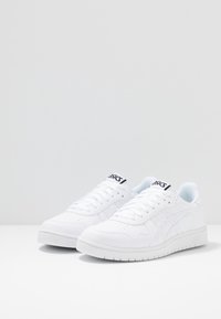 Bílé sportovní tenisky s hladkým koženým svrškem, strukturovanými detaily, perforacemi a bílou gumovou podrážkou. Na jazyku je černé logo.