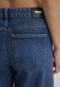 Blue denim jeans with a high waist, featuring a Mavi logo tag, distinct back pocket design, and a textured woven fabric surface.