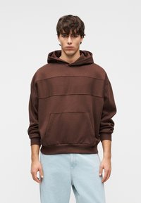 Καφέ φούτερ με κουκούλα και τσέπη μπροστά, με χαλαρή εφαρμογή και απαλό ύφασμα. Συνδυασμένο με ανοιχτόχρωμο τζιν παντελόνι.