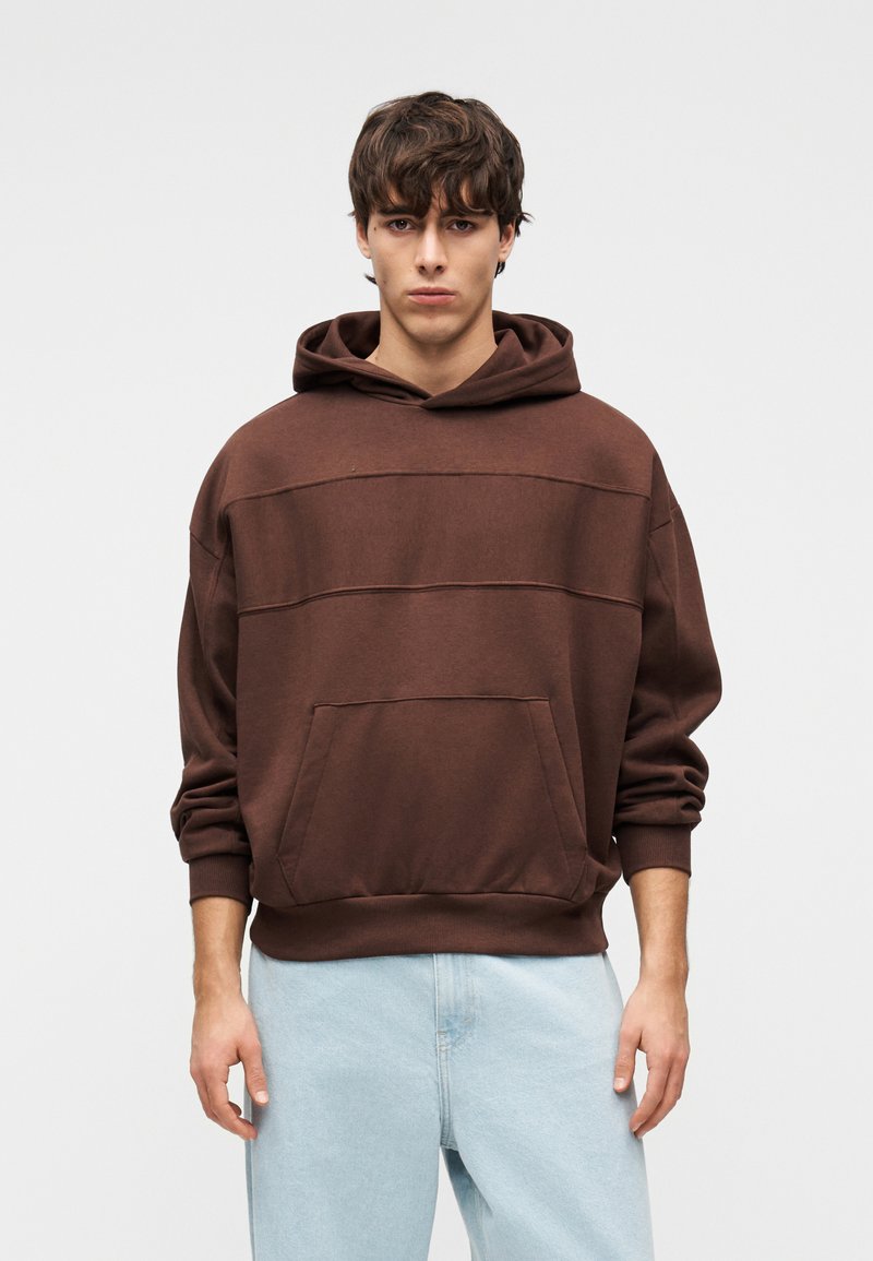 Καφέ φούτερ με κουκούλα και τσέπη μπροστά, με χαλαρή εφαρμογή και απαλό ύφασμα. Συνδυασμένο με ανοιχτόχρωμο τζιν παντελόνι.