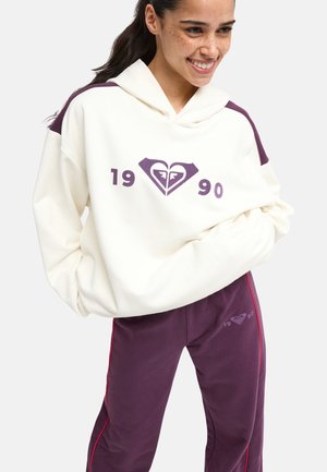 Femme souriante portant un sweat à capuche blanc avec un logo violet et des chiffres, assorti d’un pantalon violet avec un liseré rouge et un logo assorti.