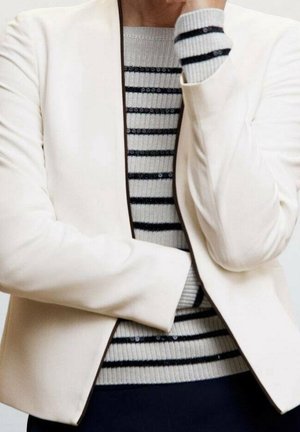 Persoon draagt een witte blazer over een lichtgrijze trui met zwarte horizontale strepen, met een arm over het lichaam gevouwen.