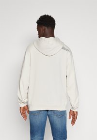 Hellgrauer Hoodie mit Kordelzug, aus weichem Material. Mit abgerundetem Saum und Textdetails auf der Rückseite. Kombiniert mit blauen Jeans.