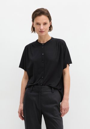 Frau mit kurzen Haaren, die eine lockere schwarze Bluse mit Knöpfen und schwarze, maßgeschneiderte Hosen vor einem schlichten hellen Hintergrund trägt.