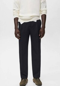 Homme portant un pantalon droit noir, un pull blanc et des chaussures lacées en daim marron, se tenant contre un fond blanc uni.