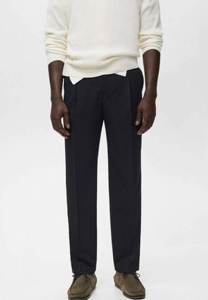 Man wearing black straight-leg pants, a white sweater, and brown suede lace-up shoes, standing against a plain white background.