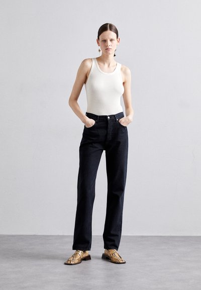 Svart denim høytalende jeans, ribbet hvit tanktop og mønstrede gullsko. Enkel design; modellen står med hendene i lommene.