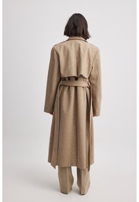 Lange beige wollen jas met een gebonden taille, grote revers en een gelaagde achterkant. Gepaard met een bijpassende broek, met een op maat gemaakte pasvorm.