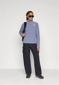 Šviesiai mėlynos spalvos džemperis su aukštu apykakle, derinamas su tamsiomis krovinių kelnėmis kišenėmis ir juoda avalyne. Aksesuarai: akiniai nuo saulės ir kuprinė.