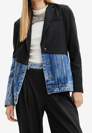 Frau trägt ein schwarzes Nadelstreifen-Blazer kombiniert mit blauen Denim-Einsätzen und schwarzen Nadelstreifen-Hosen, kombiniert mit einem weißen, gerippten Oberteil.