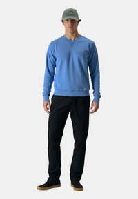 Felpa blu a maniche lunghe, con tessuto testurizzato e un emblema, abbinata a pantaloni neri tapered e scarpe slip-on nere, completata da un berretto verde.