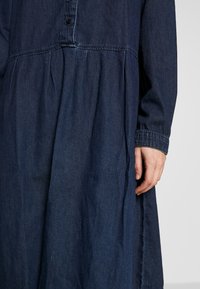 Denimklänning med mörkblått tyg, knappstängning och rynkad kjol med veck. Långa ärmar och en avslappnad passform.