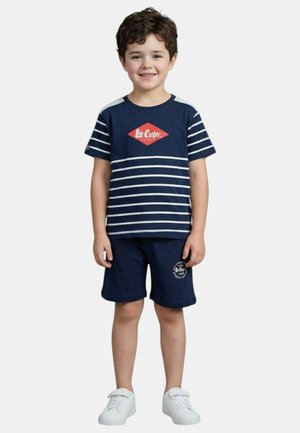 Jeune garçon souriant debout, portant un t-shirt à rayures bleu marine avec un logo rouge, un short bleu marine avec un texte blanc, et des baskets blanches.