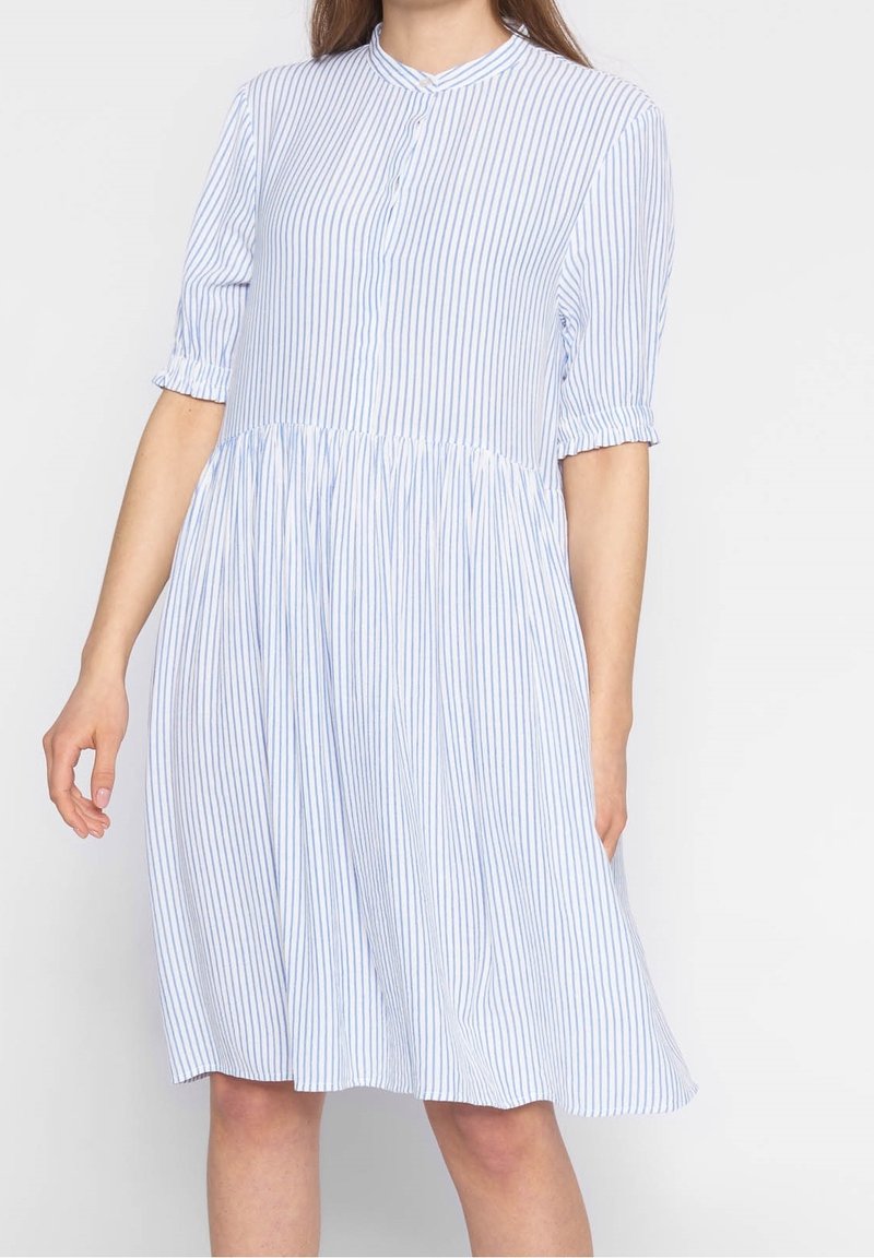 Femme portant une robe blanche ample mi-longue avec de fines rayures verticales bleues et des manches mi-longues, sur un fond uni clair.