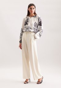 Vrouw in witte wijde broek, zwart-witte gemusterde blouse met lange mouwen en zwarte hoge hakken, staand tegen een effen achtergrond.
