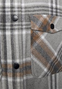 Grå rutiga skjortan är tillverkad av textil med struktur, har vita, svarta och bruna mönster, svarta knappar och en bröstfickdetalj.