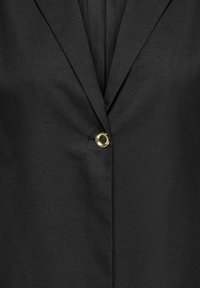 Blazer noir avec un seul bouton doré brillant attaché au centre sous les revers.