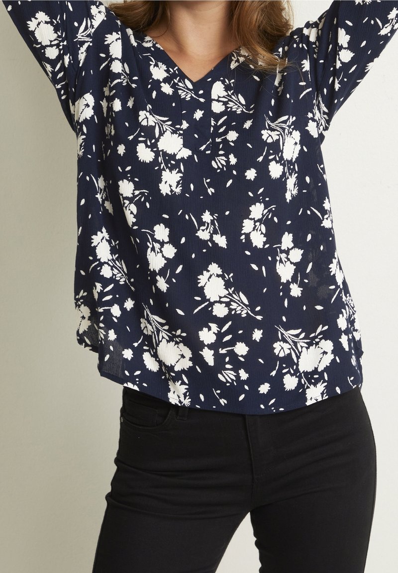 Blouse florale bleu marine avec motif de fleurs blanches, décolleté en V, manches longues et coupe décontractée. Associée à un pantalon noir ajusté.